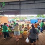 A group of people are shopping at a market, with a variety of items on display, including fruits and vegetables.