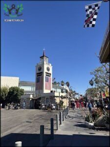 A large clock tower with a flag on top, surrounded by palm trees and people walking by.