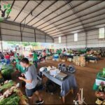 A man in a grey shirt is walking through a market filled with people and tables.