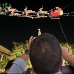 A man is taking a picture of a Christmas display featuring a Santa Claus sleigh.