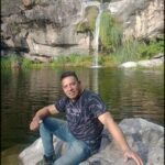 A man poses for a picture in front of a waterfall, wearing a black shirt and blue jeans.