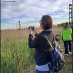 A woman in a black sweater is taking a picture of the tall grass with her cell phone.