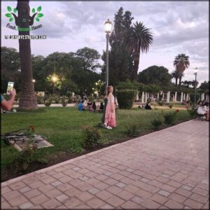 A woman in a flowered dress stands in a park, taking a selfie with her cell phone.