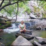 A woman poses for a picture in front of a waterfall, surrounded by rocks and trees.