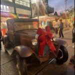 An old-fashioned car with a red bow on the front is parked on a street.