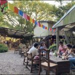 people sitting at tables outside of a restaurant with flags hanging from the roof
