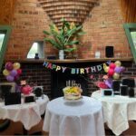 A birthday cake is placed on a table in front of a fireplace, surrounded by balloons and flowers.