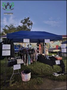 A blue tent is set up in a grassy area, with a table full of clothes and shoes underneath it.