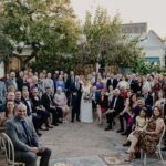 A bride and groom stand in front of their wedding party, surrounded by friends and family.