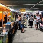 A busy street with people walking and a food stand with a man in a Hawaiian shirt standing in front of it.