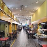 A caption for the image could be: "A long, narrow walkway in a shopping mall is illuminated by lights and features a variety of dining options, including a food court and several restaurants."