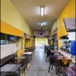 A caption for the image could be "The interior of a restaurant with a yellow and white color scheme, featuring a dining area with tables and chairs, and a kitchen area with a stove and oven."