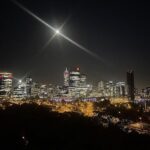 A city skyline is lit up at night, with a bright moon in the sky.