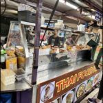 A food stand with a man standing behind it, selling Thai food.