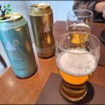 A glass of beer sits on a table next to two cans of beer.