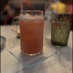 A glass of pink drink with a straw in it sits on a table next to a bottle.