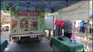 A green table is set up in front of a truck, with a sign on the table advertising organic products.
