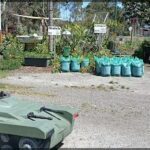 A green tank-like vehicle is parked in a dirt lot, surrounded by bags of mulch and potted plants.