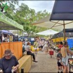 A group of people are gathered around a market, with some sitting on a bench and others standing. They are enjoying the outdoor market atmosphere and possibly shopping for fresh produce.