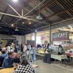 A group of people are gathered in a large building, possibly a bakery or a market, with a variety of food items on display.