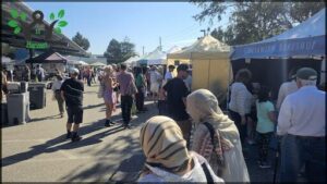 A group of people are gathered in a street, with some of them wearing headscarves. They are standing in front of a tent, possibly at a market or an event.