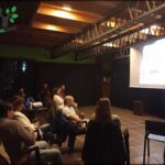A group of people are sitting in a dark room, watching a presentation on a large screen.