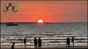 A group of people are standing on the beach, watching the sun set over the ocean.