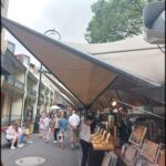 A group of people are walking down a street under a large umbrella, possibly in a market setting.