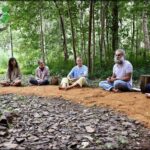 A group of people sit on the ground in a forest, listening to a man speak.