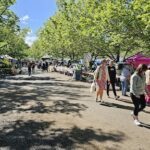 A group of people walking down a street, with a woman in a floral dress walking past a market stand.