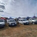 A group of white vans are parked in a field, with a cloudy sky above them.
