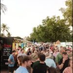 A large crowd of people gathered in a public space, with many individuals wearing hats and some carrying handbags. The atmosphere appears lively and bustling, as people mingle and enjoy the event.