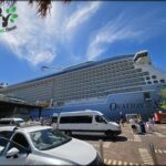 A large cruise ship is docked at a pier, with a white van and a few people nearby.