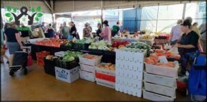 A large group of people are gathered around a market stand, browsing the fresh produce available.