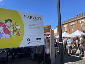 A large group of people are gathered around a sign for Harvest Farmers Market.