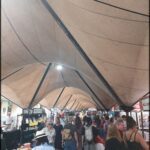 A large group of people are gathered under a tent, with some of them carrying backpacks and handbags.