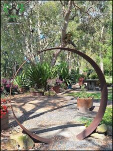 A large metal circle sits in a garden, surrounded by trees and flowers.