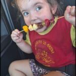 A little girl is eating fruit on a stick while sitting in a car.
