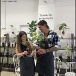 A man and woman are holding up small plants, possibly at a nursery or plant shop.