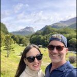 A man and woman are posing for a picture in front of a beautiful mountain range.