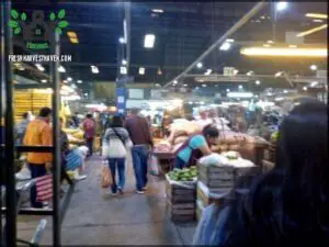 A man and woman walk through a market, passing by a table with fruits and vegetables.