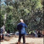 A man in a blue jacket is talking on his cell phone while standing in a park.