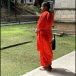 A man in an orange robe stands on a concrete ledge, possibly meditating or observing his surroundings.