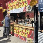 A man is standing in front of a food stand, which is selling Indonesian Martabak.