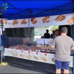 A man is standing in front of a food stand, which offers a variety of sandwiches and other food items.