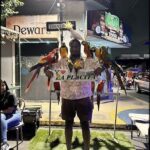 A man is standing in front of a sign that says "I love La Placita." He is surrounded by a group of colorful birds, including parrots, that are perched on his shoulders and arms.