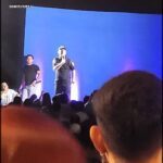 A man is standing on stage, holding a microphone, and addressing a crowd of people.