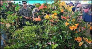 A man stands among a variety of colorful flowers, including red, yellow, and orange ones.