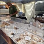A man stands behind a table filled with various pastries, including donuts and bread.