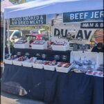 A man stands behind a table filled with various types of meat, including ham and beef jerky.